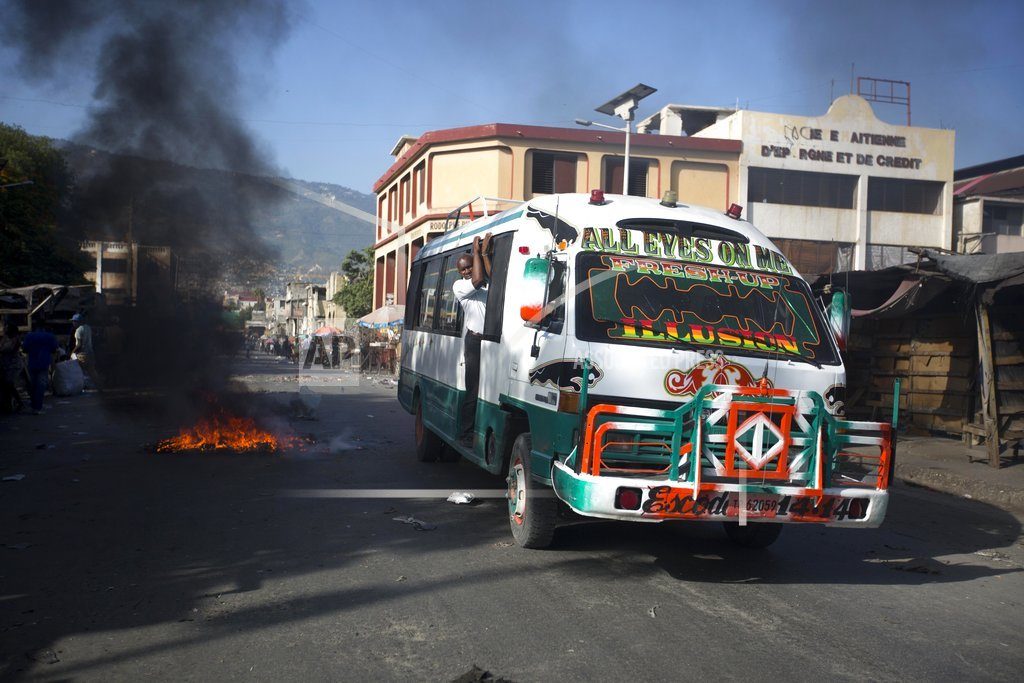 Un camión lleno de pasajeros pasa junto a llantas incendiándose durante una huelga que demanda la renuncia del presidente Jovenel Moise en Puerto Príncipe, Haití, el lunes 30 de julio de 2018. ( AP Foto/Dieu Nalio Chery)